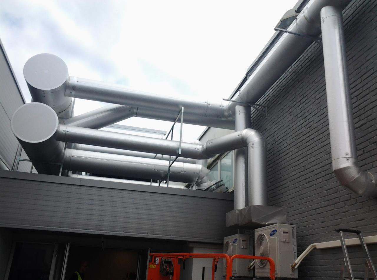 Srebrne rury wentylacyjne na zewnątrz budynku, z widocznymi jednostkami klimatyzacji i podnośnikiem koszowym w kolorze pomarańczowym.