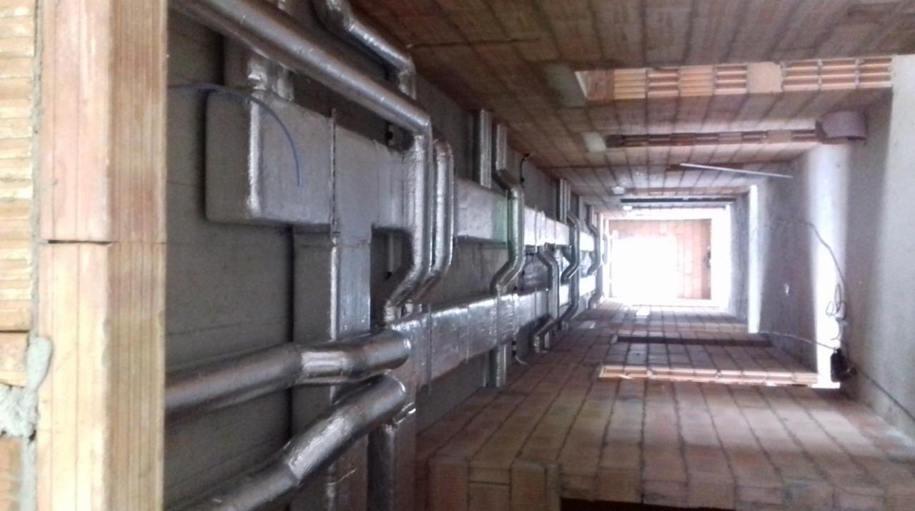 Wnętrze budynku w trakcie montażu instalacji wentylacyjnej z widocznymi izolowanymi kanałami wentylacyjnymi biegnącymi wzdłuż ściany i pod sufitem, ceglana konstrukcja sufitu i podłogi.