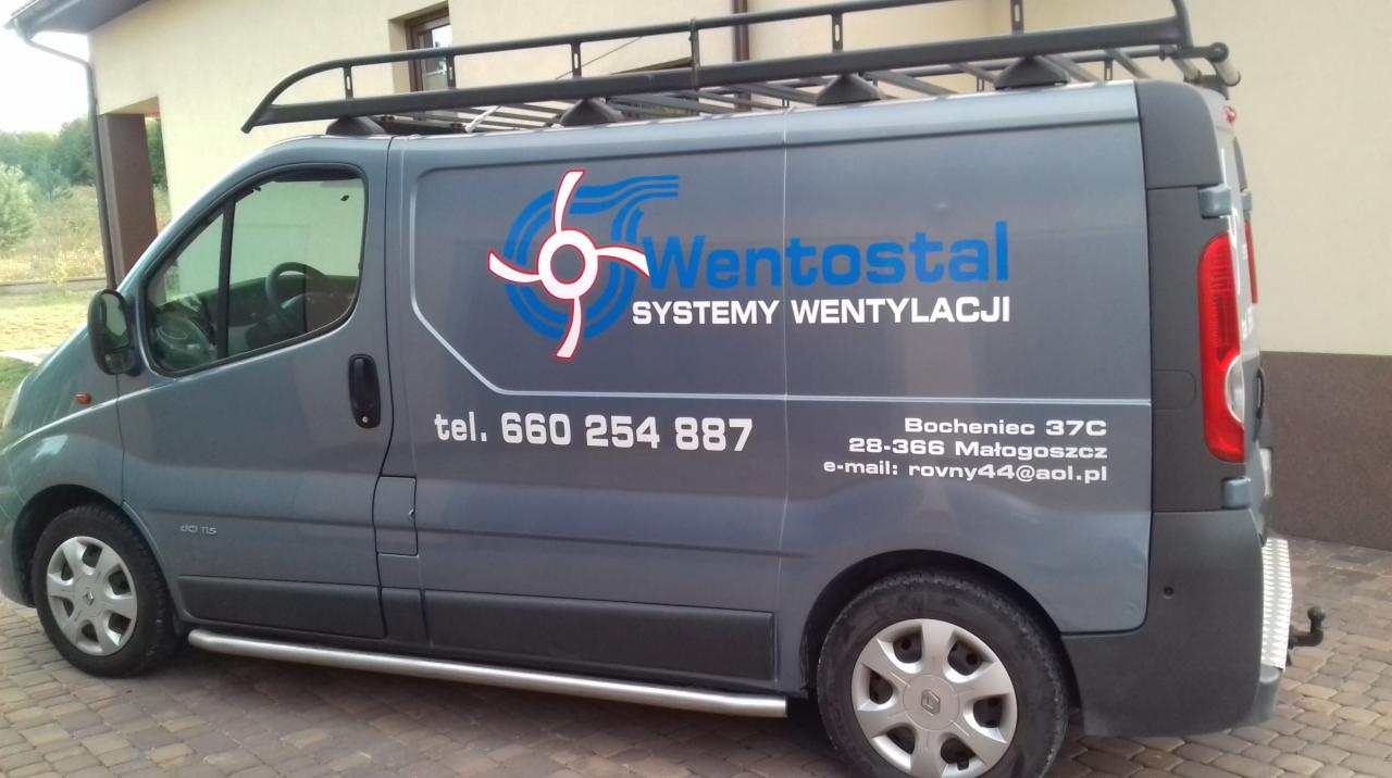 Szary samochód dostawczy z logo firmy Wentostal Systemy Wentylacji, numerem telefonu i adresem e-mail na boku, zaparkowany na zewnątrz budynku.