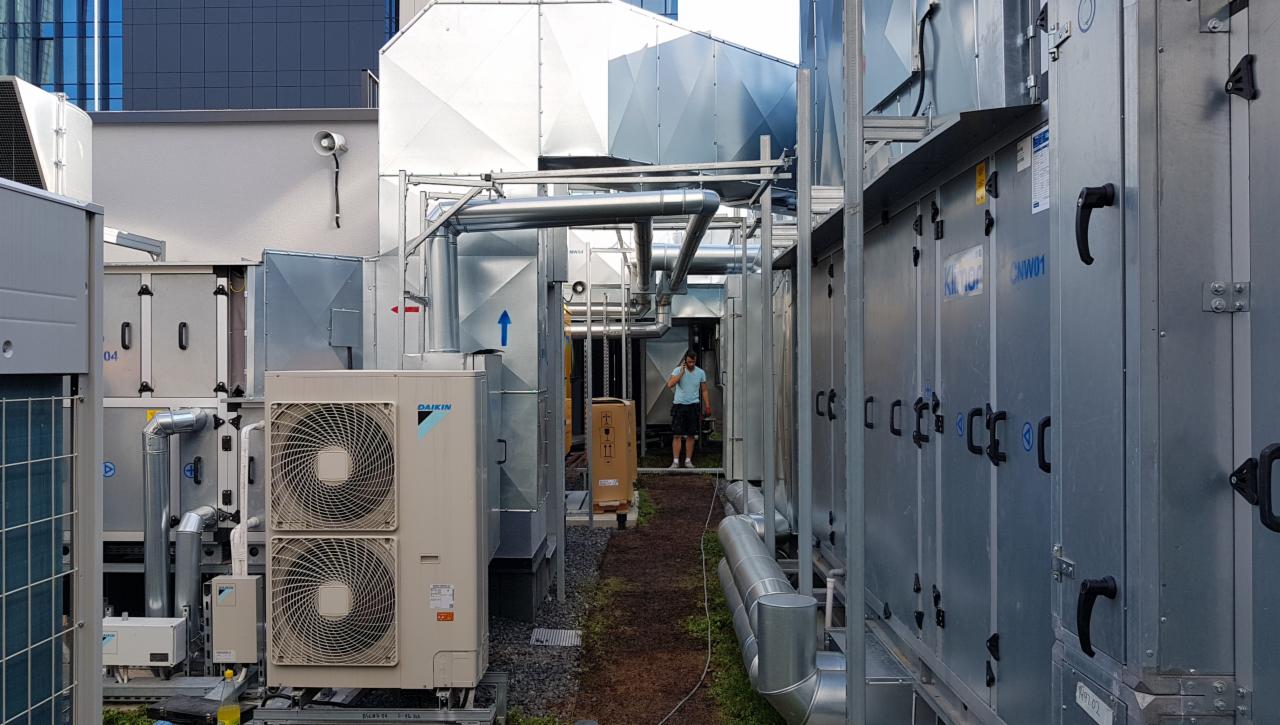 Instalacja wentylacyjna na dachu budynku, widoczny agregat Daikin, rury wentylacyjne, centrala wentylacyjna, pracownik w tle rozmawia przez telefon.