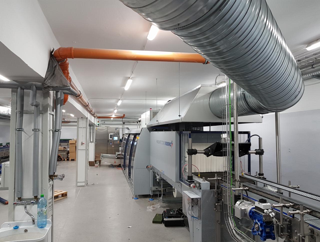 Wnętrze hali przemysłowej z zainstalowaną rozbudowaną instalacją wentylacyjną z rurami spiro i pomarańczowymi rurami PCV pod sufitem, nad linią produkcyjną z automatycznym owijaniem palet.