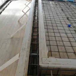 Zbrojenie pionowe w drewnianym szalunku z widoczną siatką zbrojeniową na drewnianym podłożu, niebieskie elementy styropianu przytwierdzone do zbrojenia.