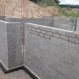 Wznoszone ściany fundamentowe z bloczków betonowych z wystającymi prętami zbrojeniowymi, zabezpieczone czarną izolacją przeciwwilgociową u podstawy, widoczne narzędzia murarskie na górnej krawędzi...
