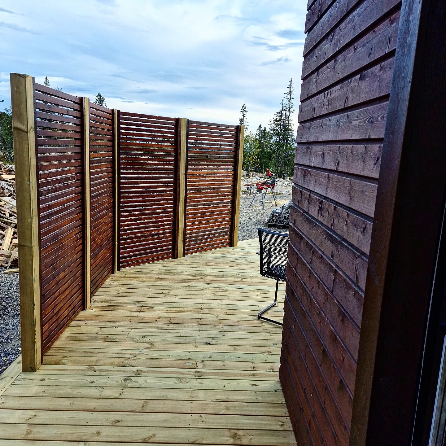 Nowa drewniana elewacja budynku z deskami ułożonymi poziomo, przylegająca do tarasu z jasnego drewna oraz ażurowe, ciemnobrązowe panele ogrodzeniowe z widocznym w tle krajobrazem i pracującą osobą.