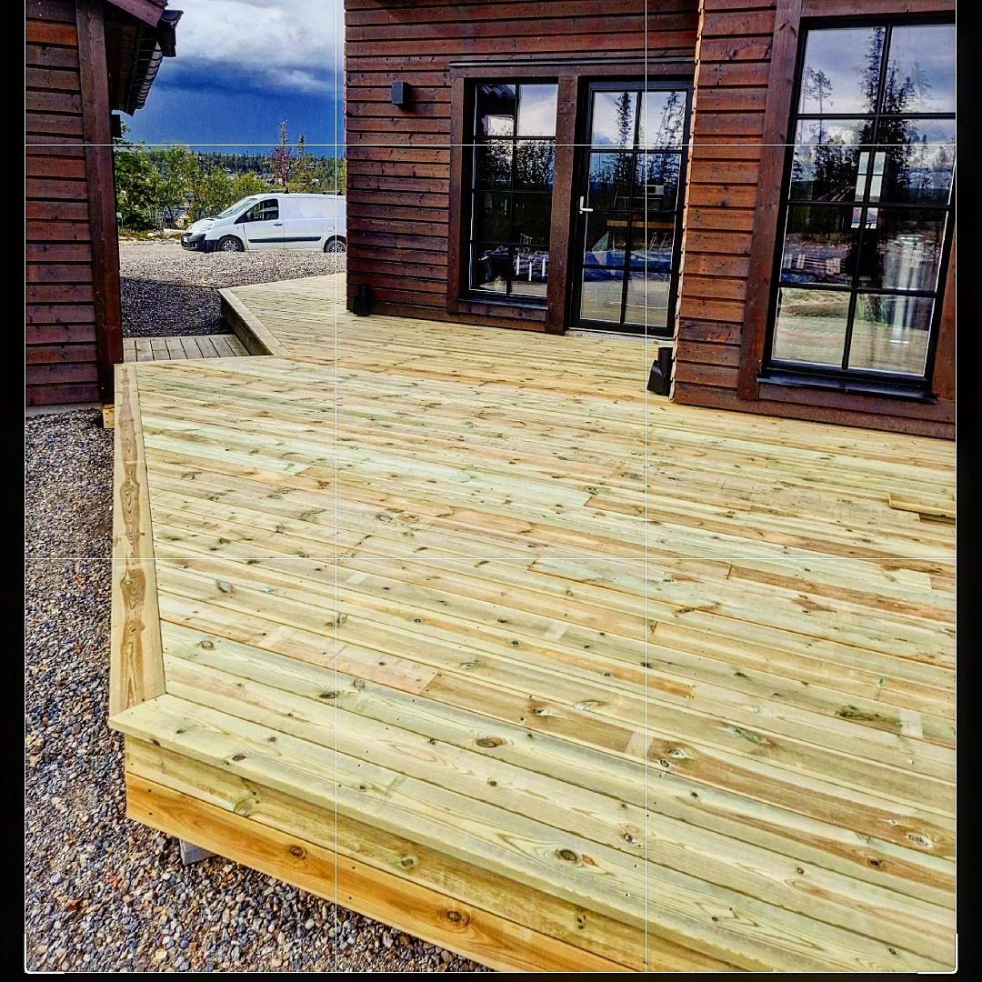 Nowo wybudowany drewniany taras przylegający do ciemnobrązowego domu z dużymi oknami, widoczny biały samochód dostawczy w tle.