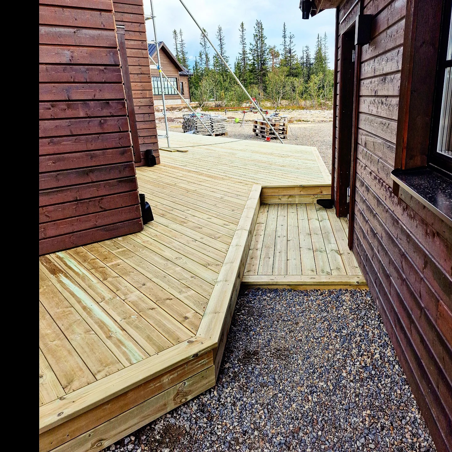 Nowo wybudowany drewniany taras z jasnego drewna, przylegający do ciemnobrązowego domu z drewnianą elewacją, widok z poziomu gruntu na żwirowe podłoże.