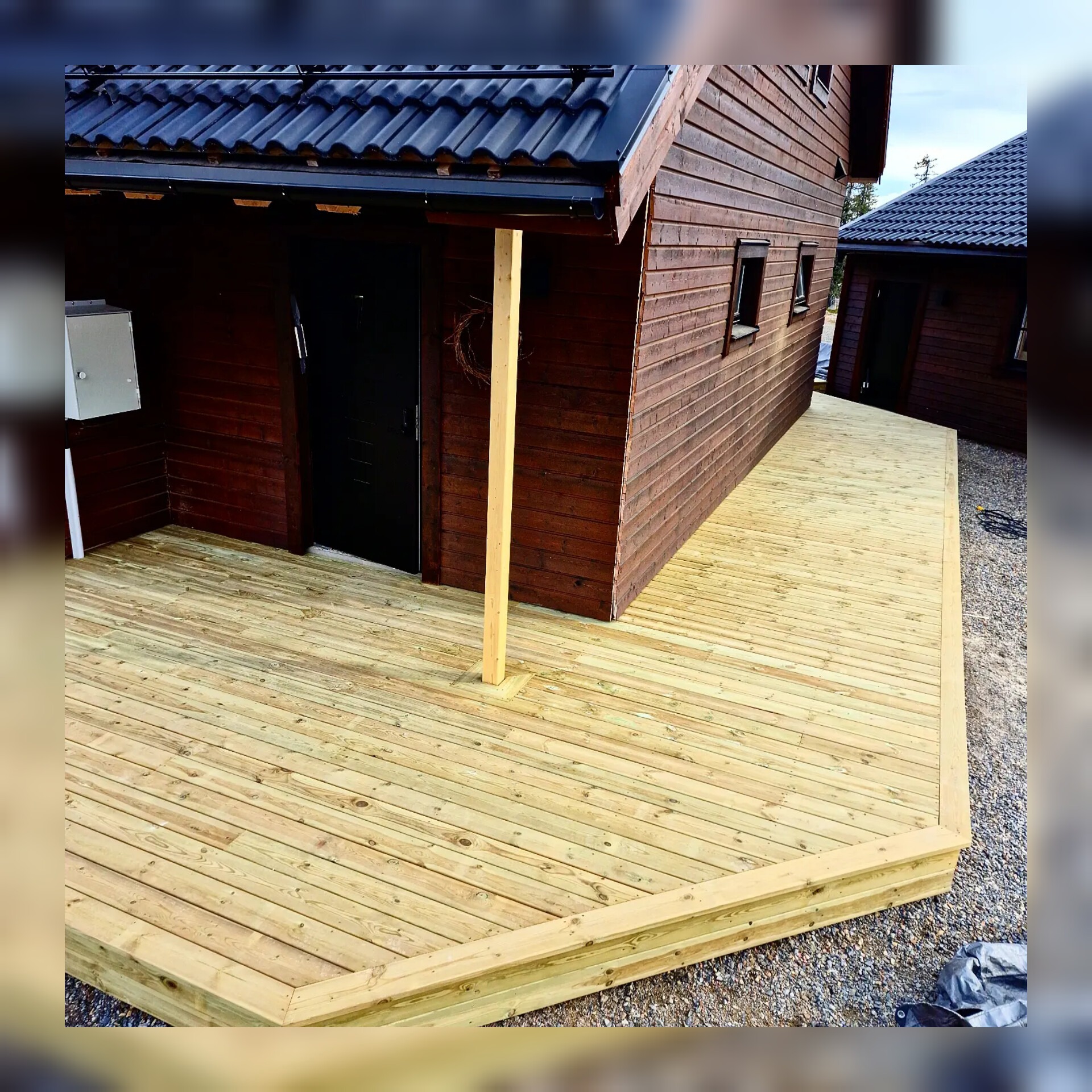 Nowo wybudowany drewniany taras przylegający do brązowego domu z ciemnym dachem, widoczna drewniana kolumna podtrzymująca zadaszenie wejścia i żwirowe podłoże.