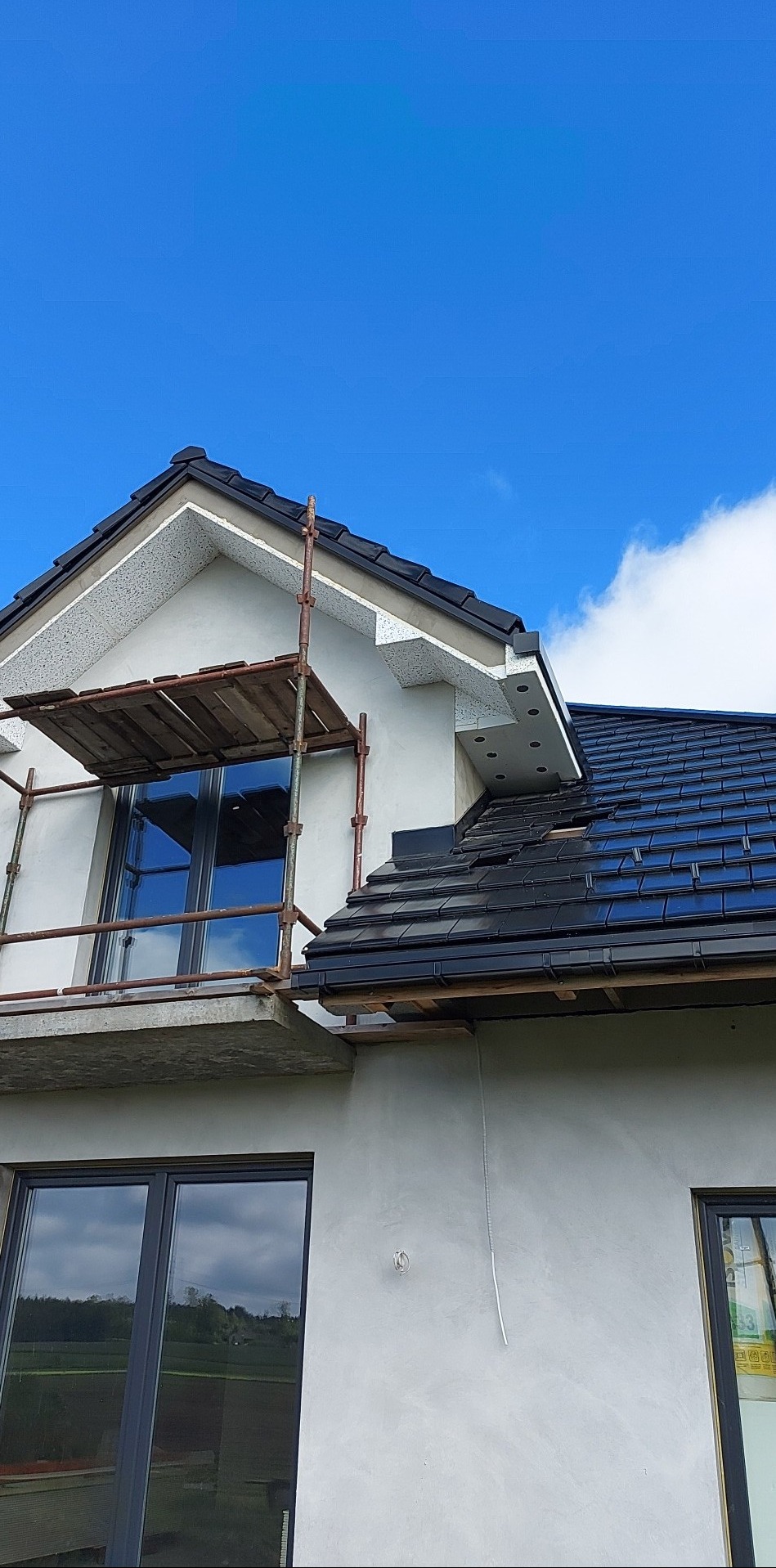 Fragment domu w Borczu w trakcie ocieplania, widoczne rusztowanie, okno z odbiciem nieba i fragment dachu z czarną dachówką.