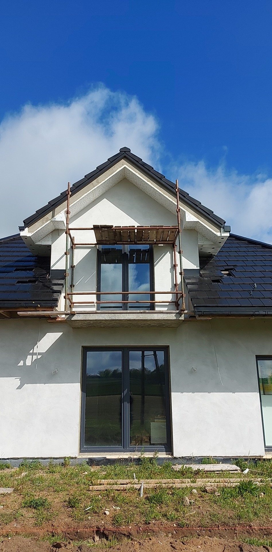 Surowy stan domu z czarną dachówką i białym tynkiem, widoczne rusztowanie przy oknie, odblask nieba w szybach okiennych.