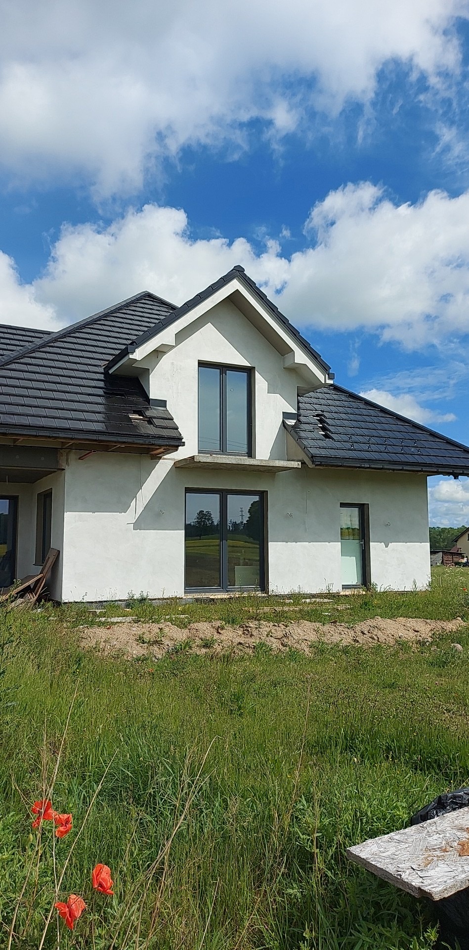 Surowy stan domu jednorodzinnego z ciemną dachówką i szarą elewacją, widoczne okna z ciemnymi ramami, w tle błękitne niebo z chmurami, na pierwszym planie trawa i czerwone maki.