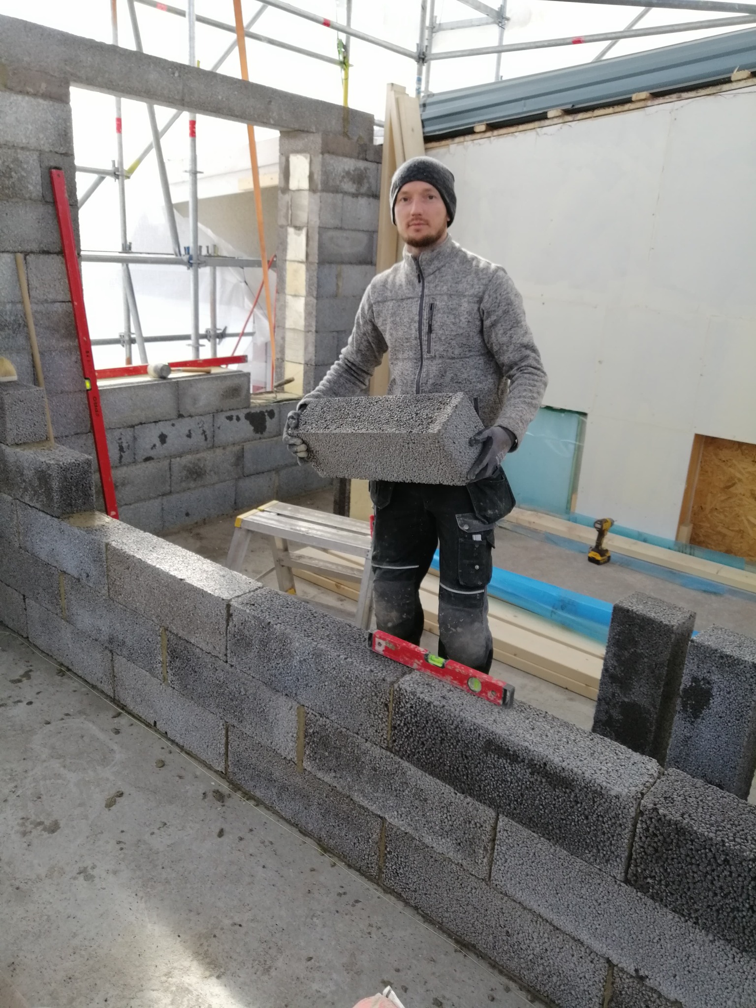 Mężczyzna w czapce i roboczych spodniach układa szare bloczki betonowe, w tle widoczne rusztowanie i narzędzia, poziomica na murze.