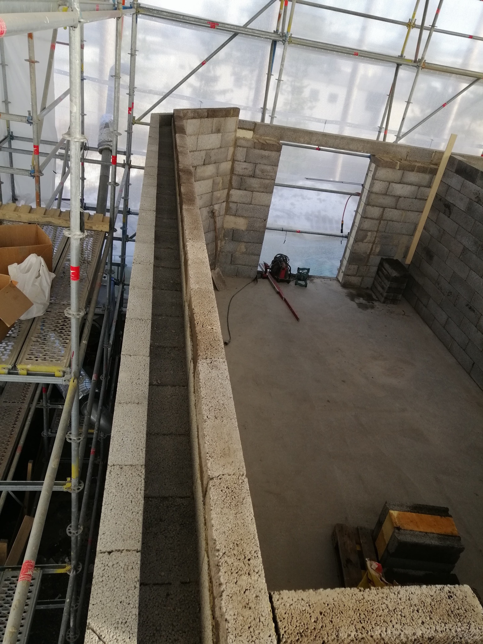 Wnętrze budynku w trakcie budowy z widocznymi ścianami z betonowych bloczków, rusztowaniami, narzędziami i materiałami budowlanymi, w tym poziomica i agregat prądotwórczy.