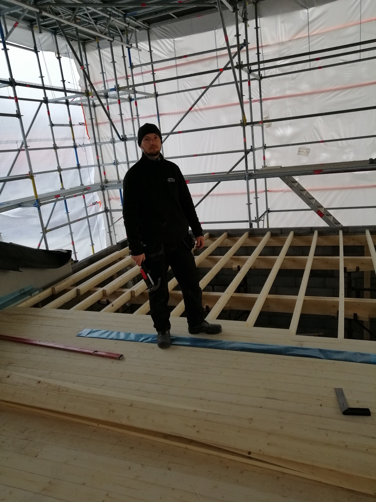 Pracownik w czarnym ubraniu i czapce, z młotkiem w ręku, stoi na drewnianej konstrukcji dachu pod rusztowaniem, w tle biała folia ochronna.