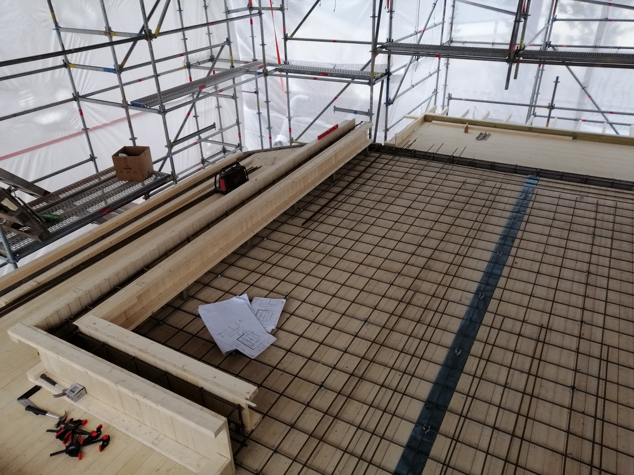 Widok z góry na szkielet zbrojeniowy ułożony na drewnianej podłodze w trakcie budowy domu, z widocznymi elementami szalunku, rusztowaniem i narzędziami.