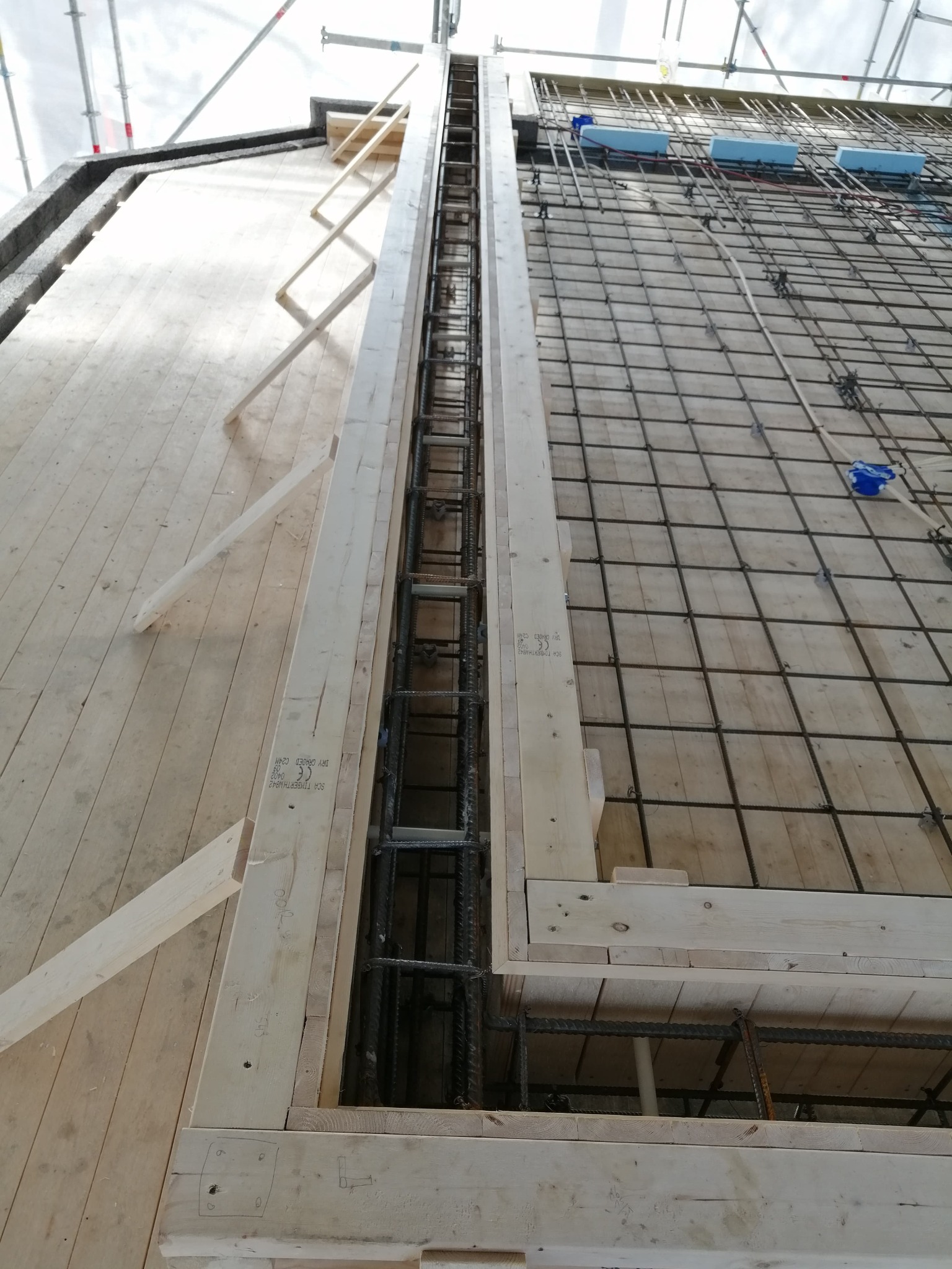 Zbrojenie pionowe w drewnianym szalunku z widoczną siatką zbrojeniową na drewnianym podłożu, niebieskie elementy styropianu przytwierdzone do zbrojenia.