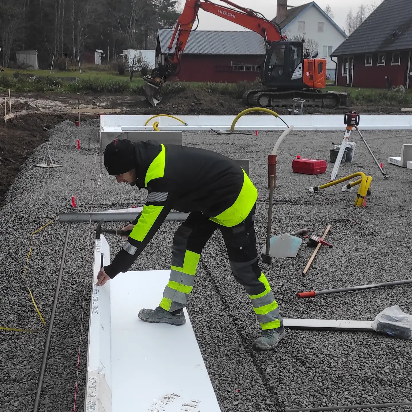 Pracownik w odzieży roboczej wbija kołek w białą płytę styropianową na podłożu z tłucznia, przygotowując fundament pod budowę, w tle koparka i budynki.