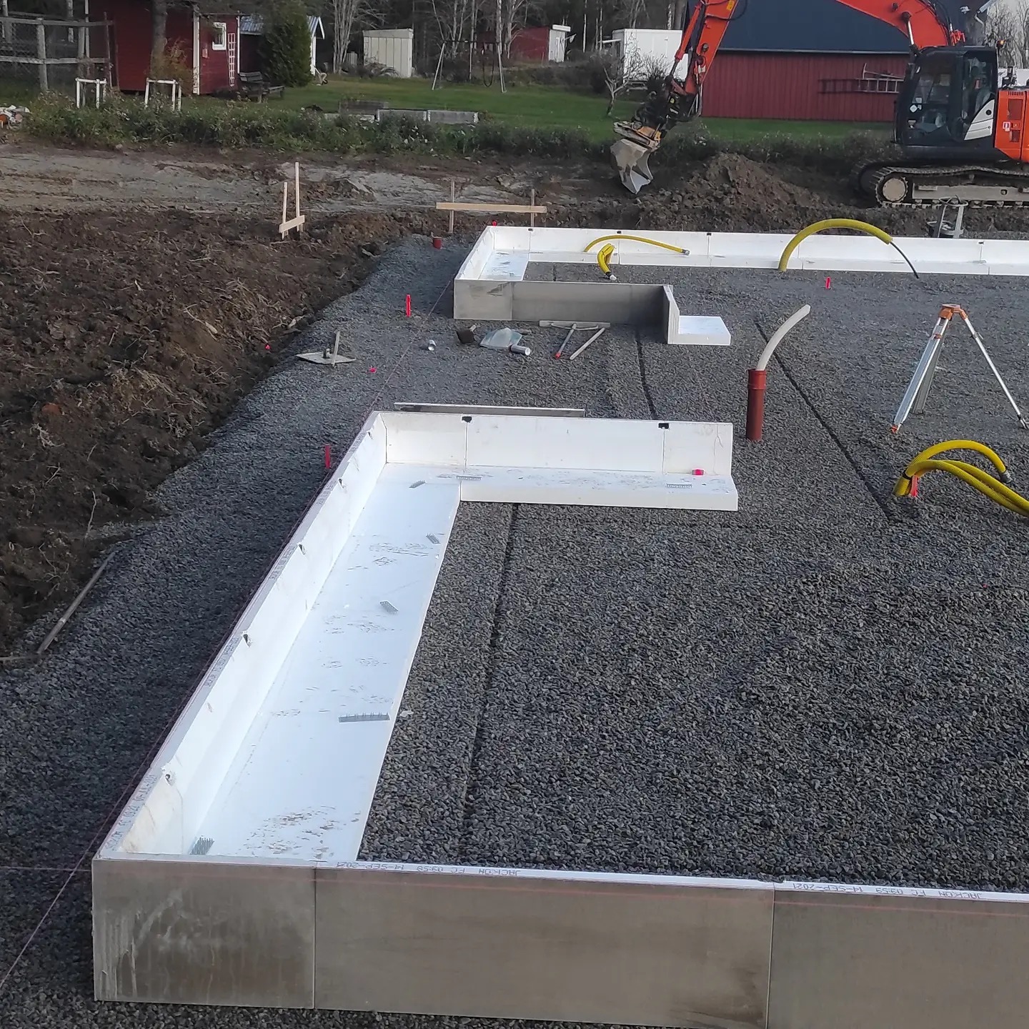 Przygotowanie fundamentów pod budowę domu: ułożone szalunki z izolacją termiczną na warstwie żwiru, koparka w tle.