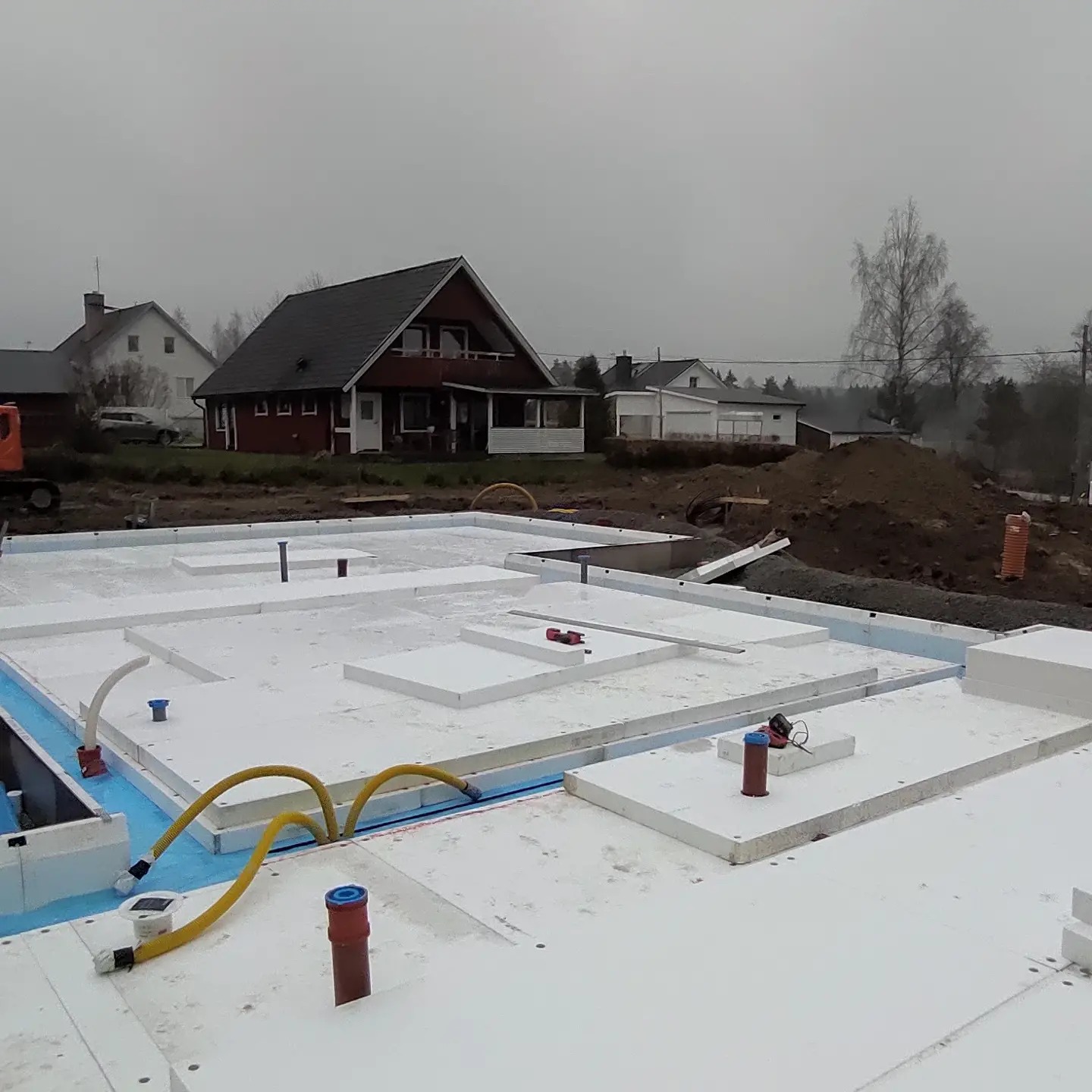 Fundament domu w trakcie budowy: białe płyty izolacyjne z widocznymi rurami instalacyjnymi, na tle domów jednorodzinnych w pochmurny dzień.