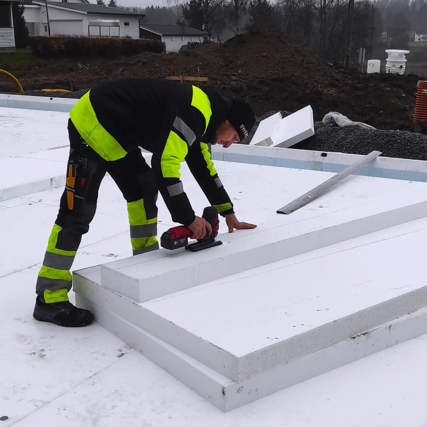 Pracownik w odzieży roboczej docina elektryczną piłą białe panele izolacyjne, prawdopodobnie styropian, na budowie domu.