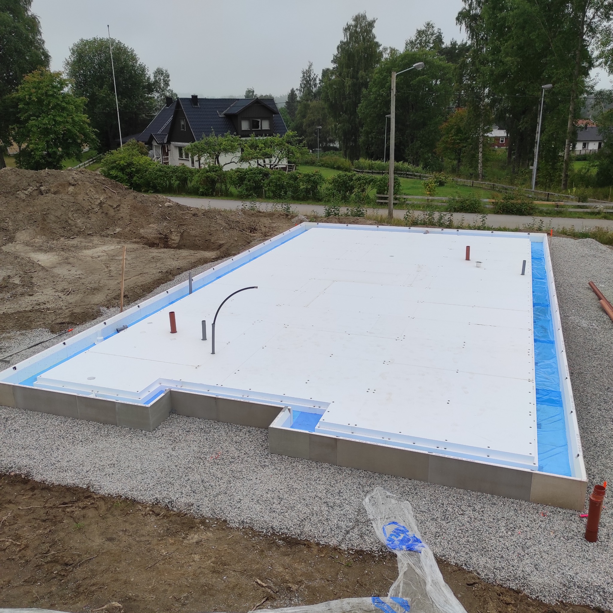 Fundament domu w trakcie budowy, widoczna płyta fundamentowa z izolacją, wystające rury instalacyjne, otoczenie z ziemi i gruzu.