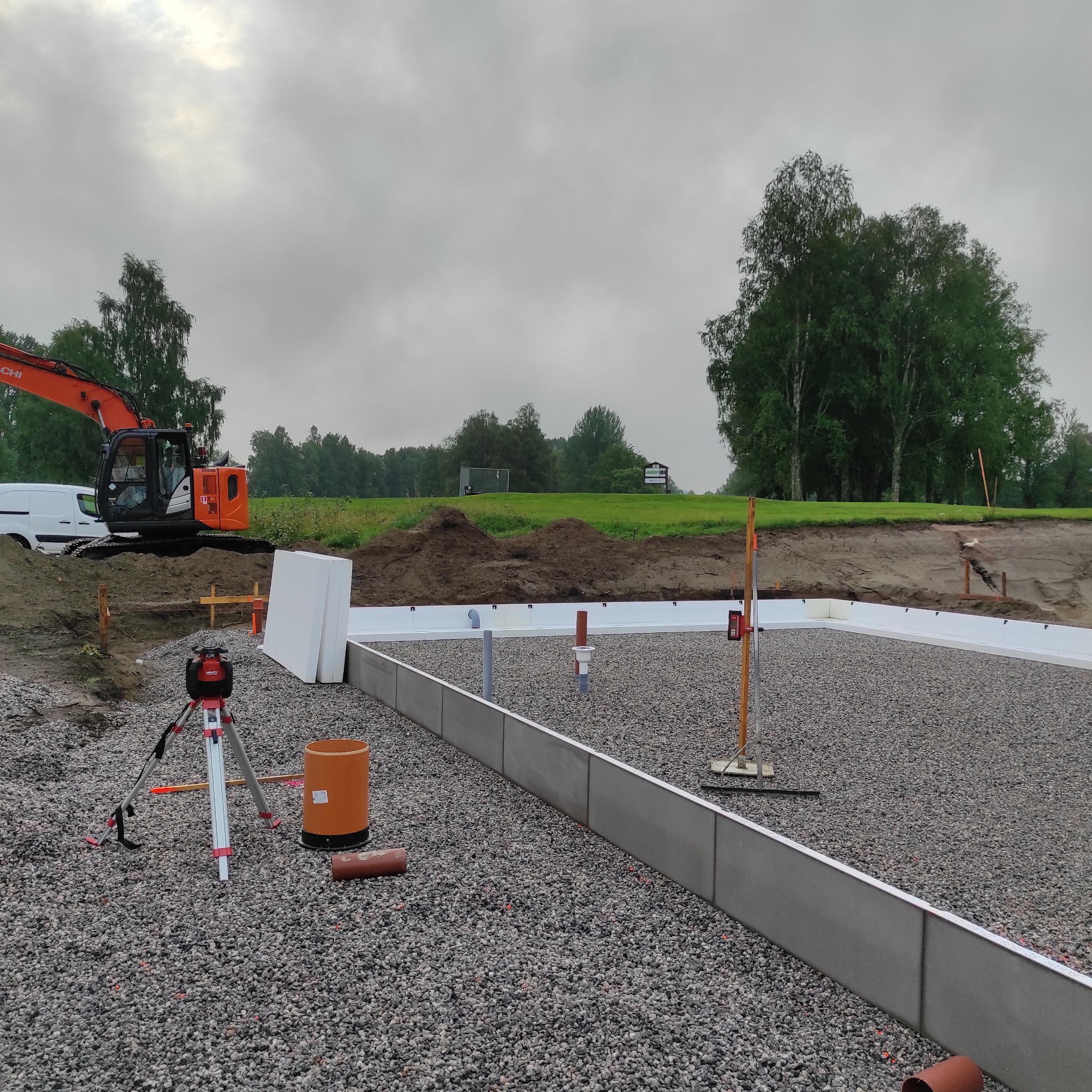 Przygotowanie fundamentów pod dom murowany: widoczny wykop z podsypką żwirową, szalunki, izolacja termiczna, instalacje pionowe i niwelator laserowy na trójnogu, w tle koparka Hitachi.