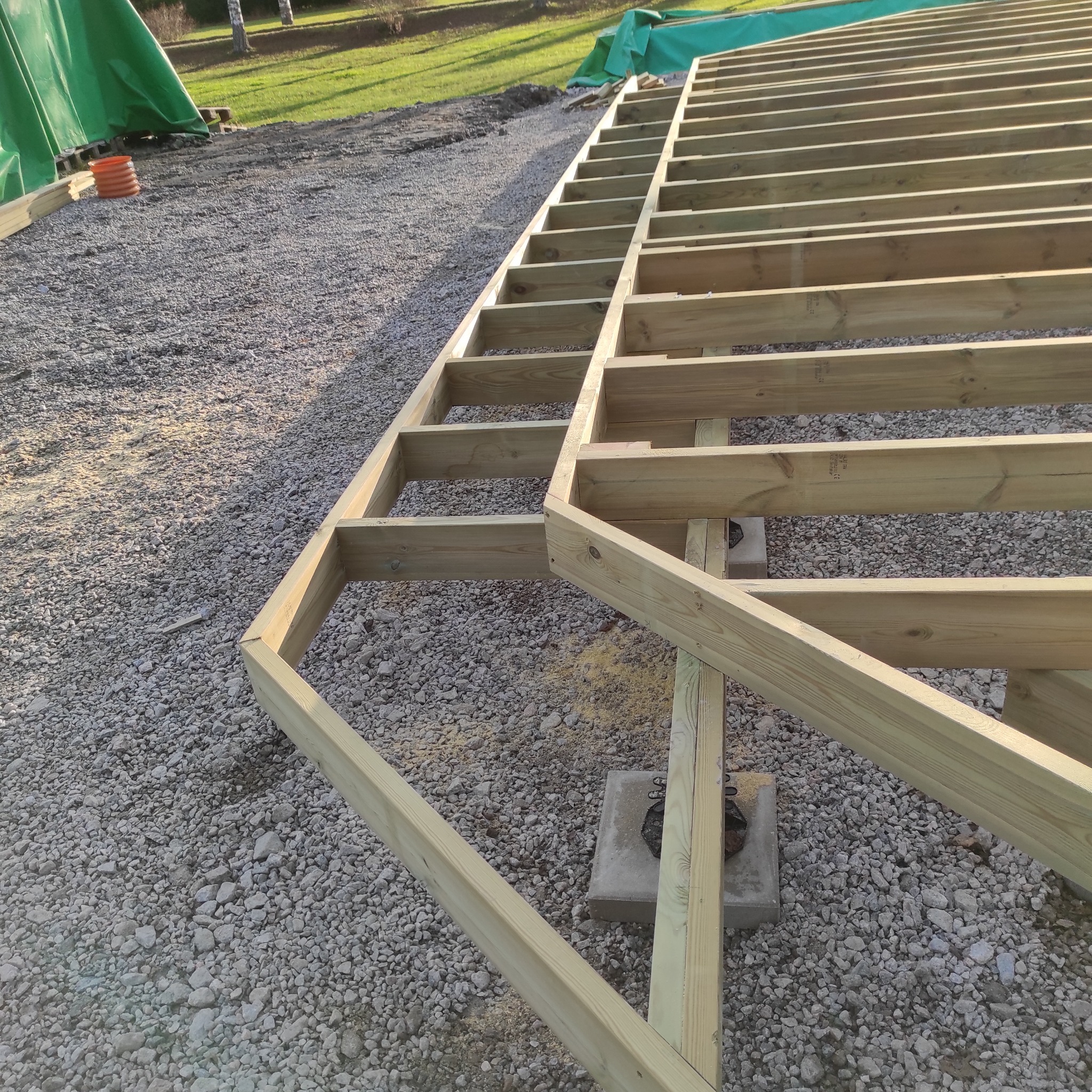 Konstrukcja drewniana pod podłogę tarasu, ułożona na betonowych bloczkach, widoczna na żwirowym podłożu z zielonymi plandekami w tle.