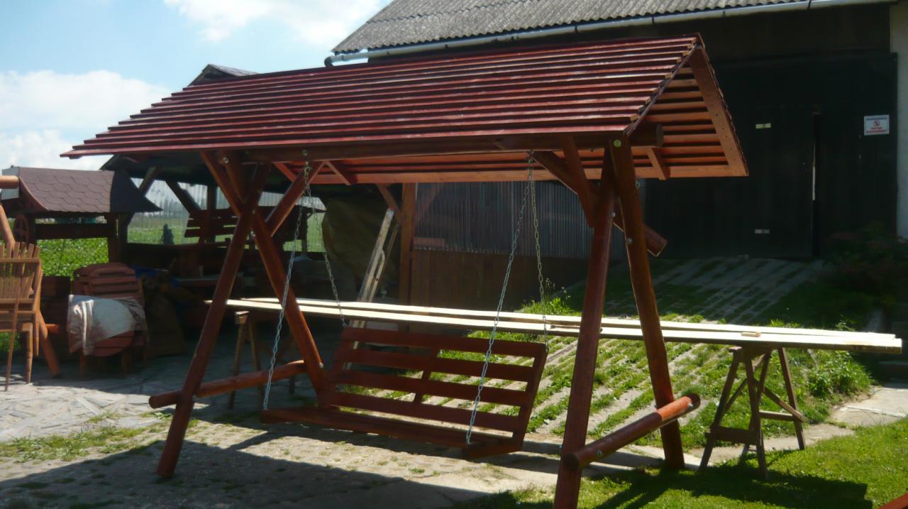 Drewniana huśtawka ogrodowa z zadaszeniem w trakcie montażu, deski na kozłach obok, w tle budynek gospodarczy.