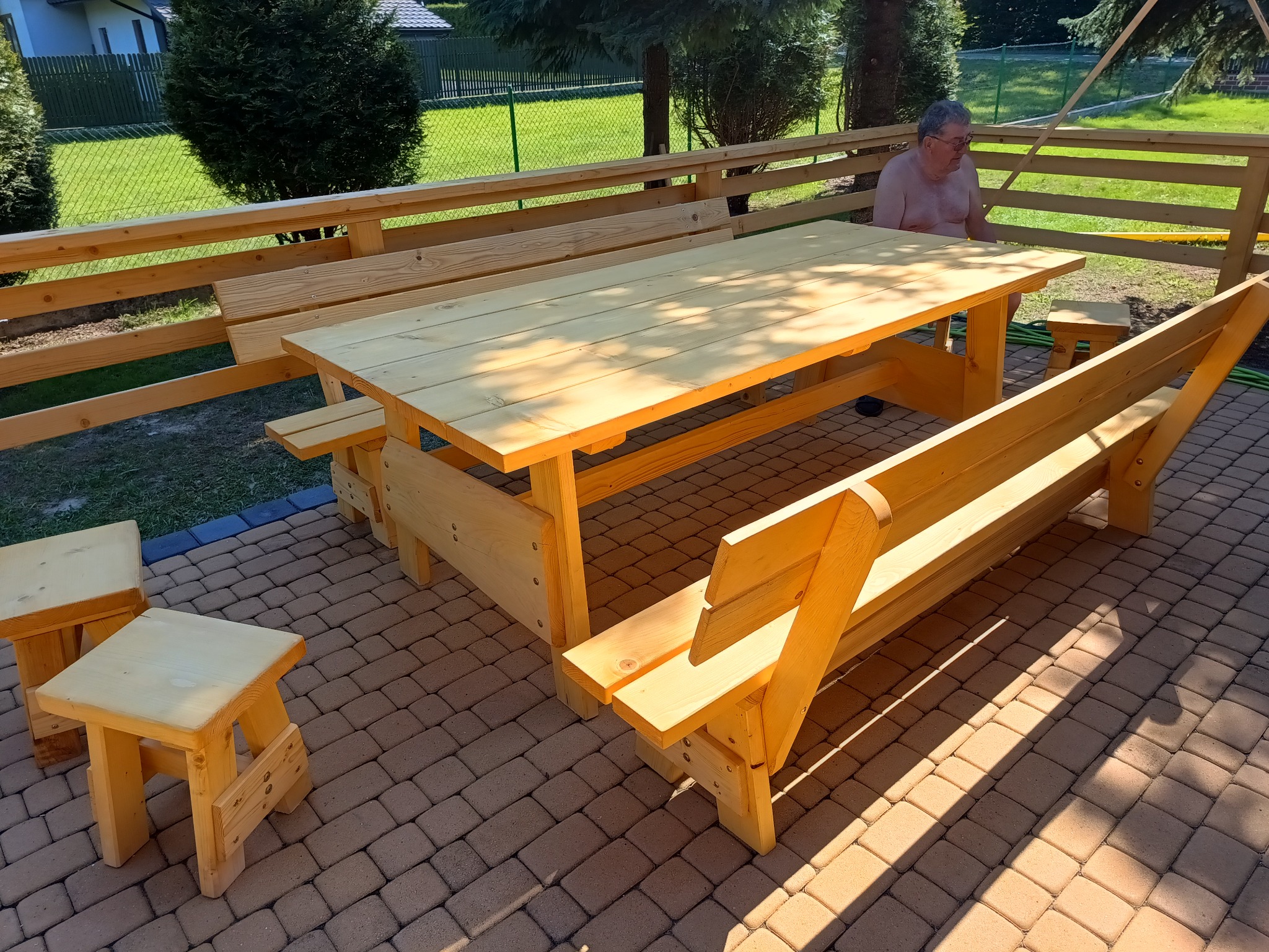 Jasne drewniane meble ogrodowe: stół z ławkami i taboretami, na brukowanym tarasie, z widocznym fragmentem ogrodzenia i siedzącym mężczyzną w tle.