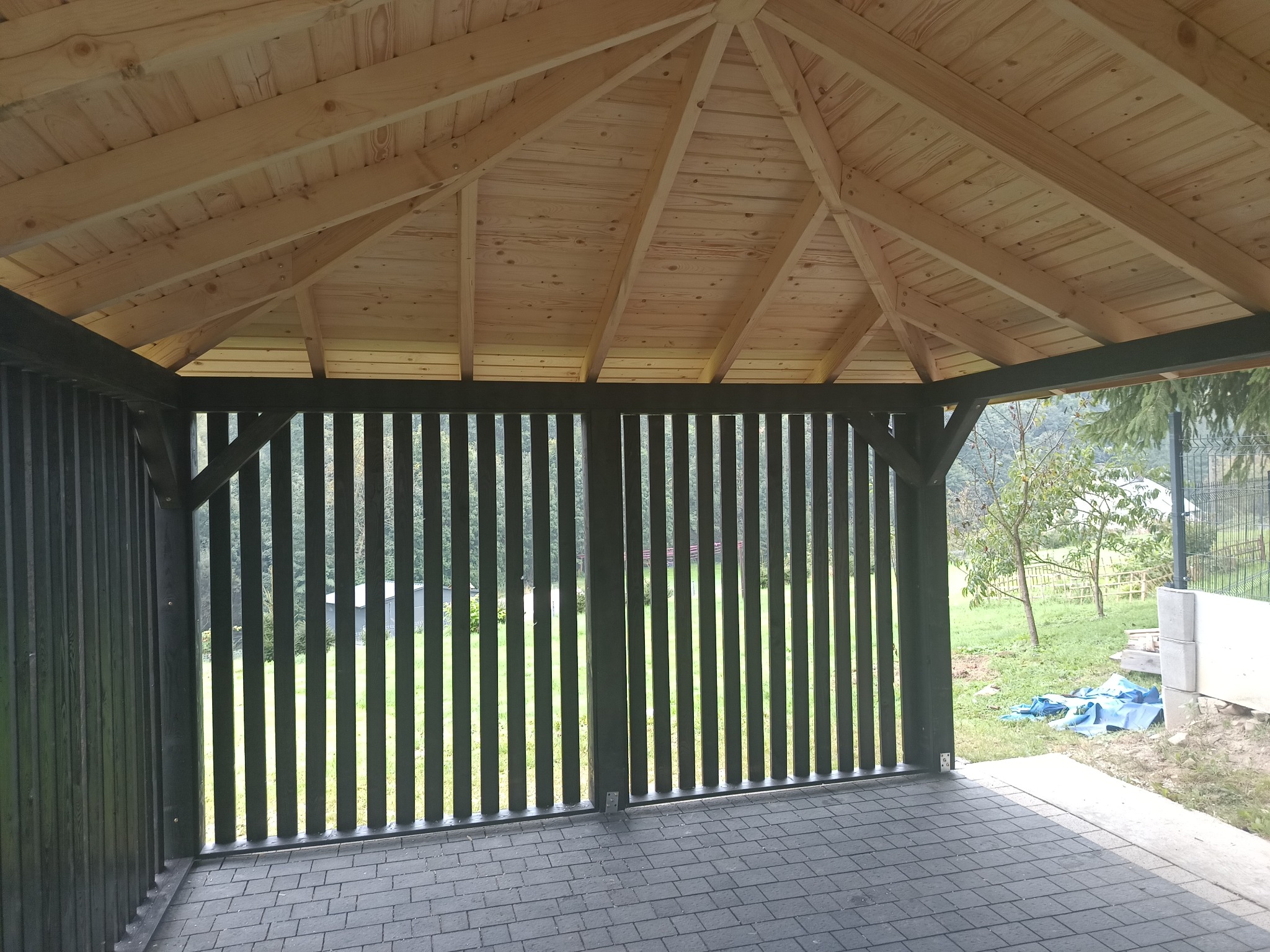 Drewniana konstrukcja altany z czarnymi pionowymi listwami, widok z wnętrza na zewnątrz, z widoczną podłogą z szarej kostki brukowej i fragmentem ogrodu w tle.