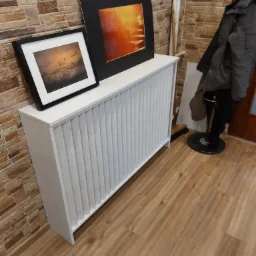 Biała obudowa grzejnika z pionowymi żłobieniami, na niej dwie oprawione grafiki, w tle ściana z cegieł i wieszak na ubrania.