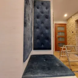 Narożne siedzisko z tapicerowanymi panelami ściennymi w odcieniach szarości, wkomponowane w białą zabudowę meblową; w tle widoczna drabina i fragment ceglanej ściany.