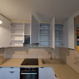 Nowoczesna kuchnia z białymi szafkami, płytą indukcyjną i piekarnikiem w zabudowie, widok na otwarte półki i dekoracyjny kalendarz na ścianie.