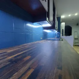 Nowoczesna kuchnia z ciemnym blatem drewnianym, niebieskimi płytkami ściennymi z delikatnym wzorem i białymi szafkami wiszącymi podświetlonymi listwą LED.