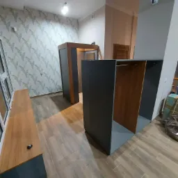 Wnętrze z częściowo zamontowaną garderobą z szarymi elementami zewnętrznymi i drewnianymi wstawkami, widoczny również fragment drewnianego blatu pod oknem i podłoga imitująca drewno.