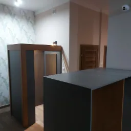 Nowoczesna zabudowa wnęki z szarą szafą i drewnianym wykończeniem, widoczna na tle ściany z wzorem liści i drewnianych drzwi.