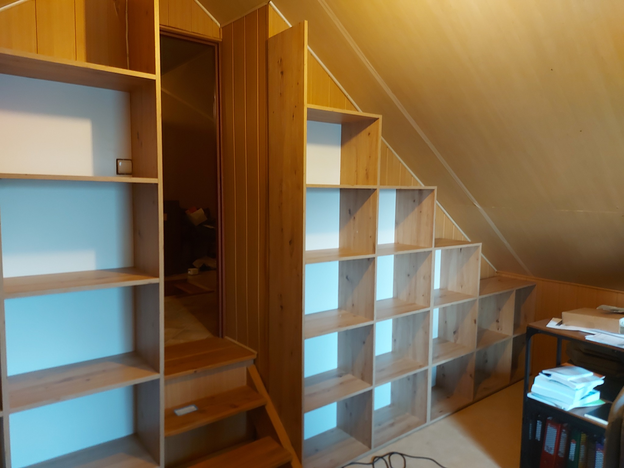 Drewniana zabudowa pod skosem na poddaszu z otwartymi półkami, zintegrowana ze schodami i szafką z oświetleniem w tle.