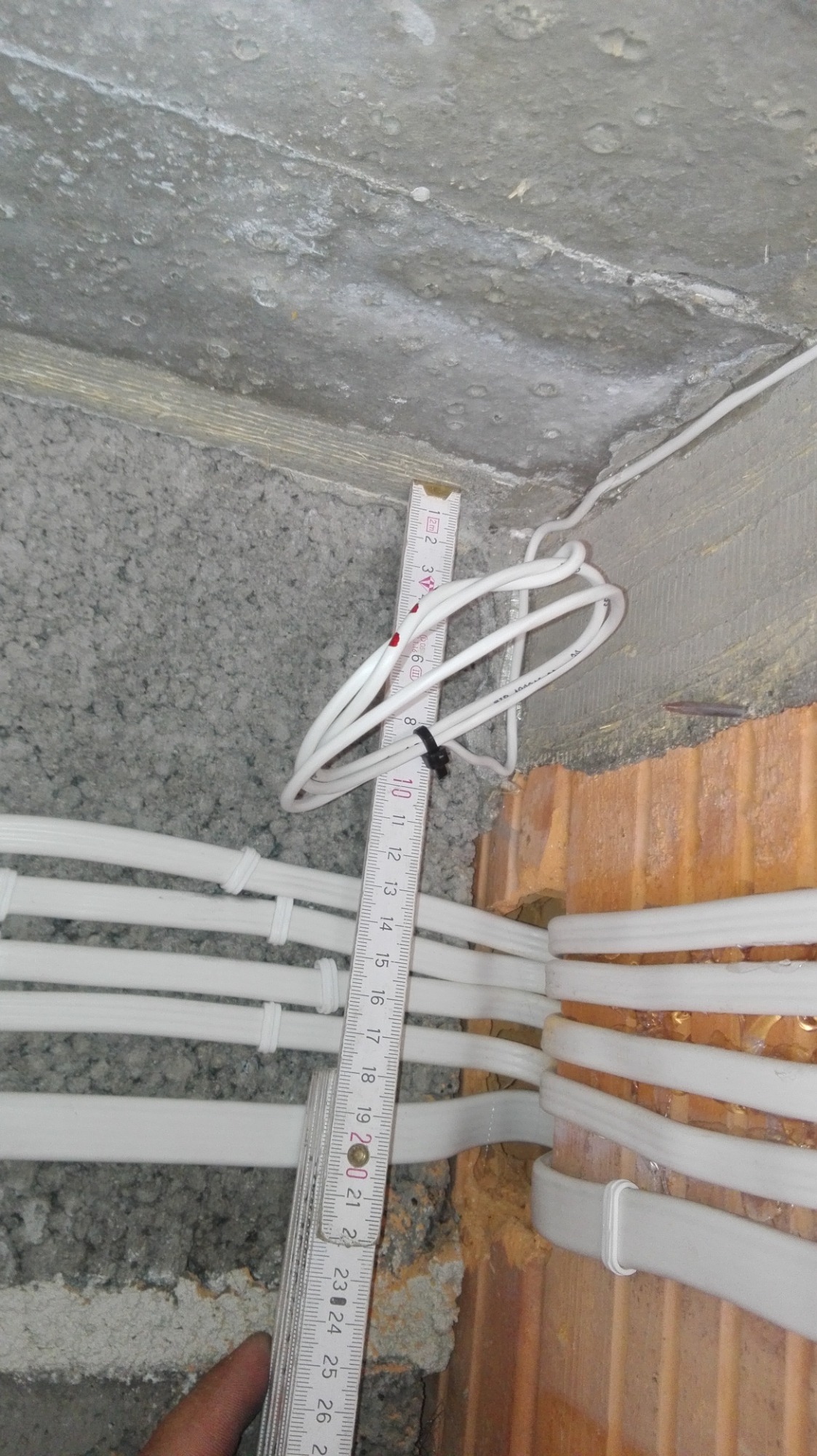 Białe kable elektryczne prowadzone wzdłuż szorstkiej, betonowej ściany, częściowo w rurkach ochronnych, mierzone rozkładaną miarką z czarnym zaciskiem na jednym z kabli.