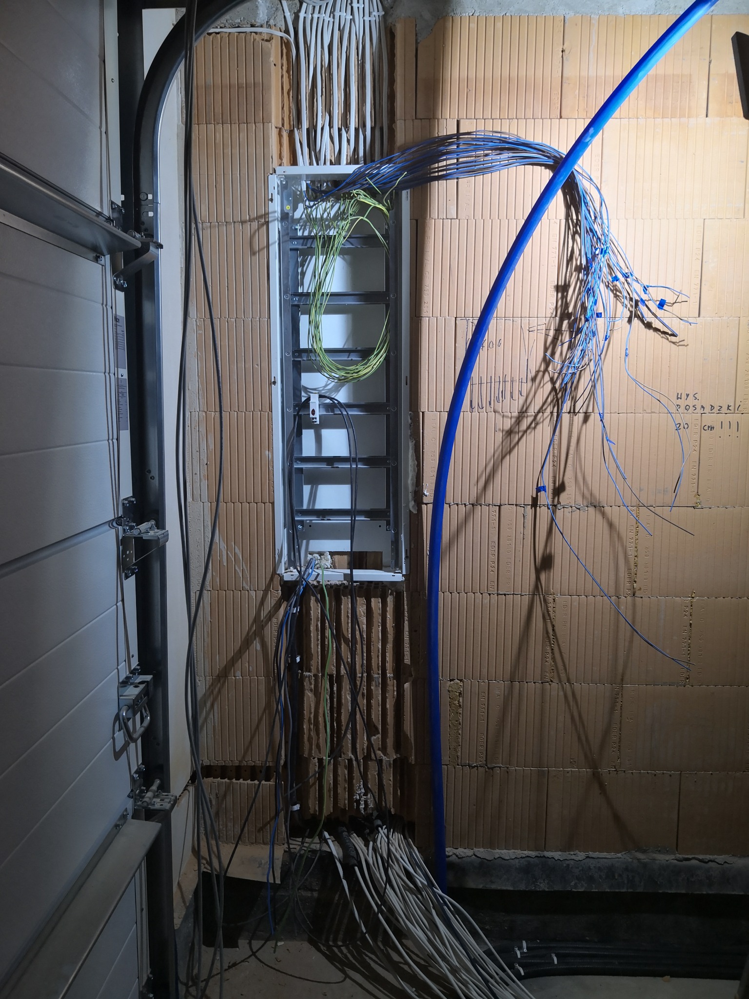 Rozdzielnica elektryczna w surowym stanie, widoczne liczne przewody, niebieska rura PCV i ceglana ściana z zapiskami