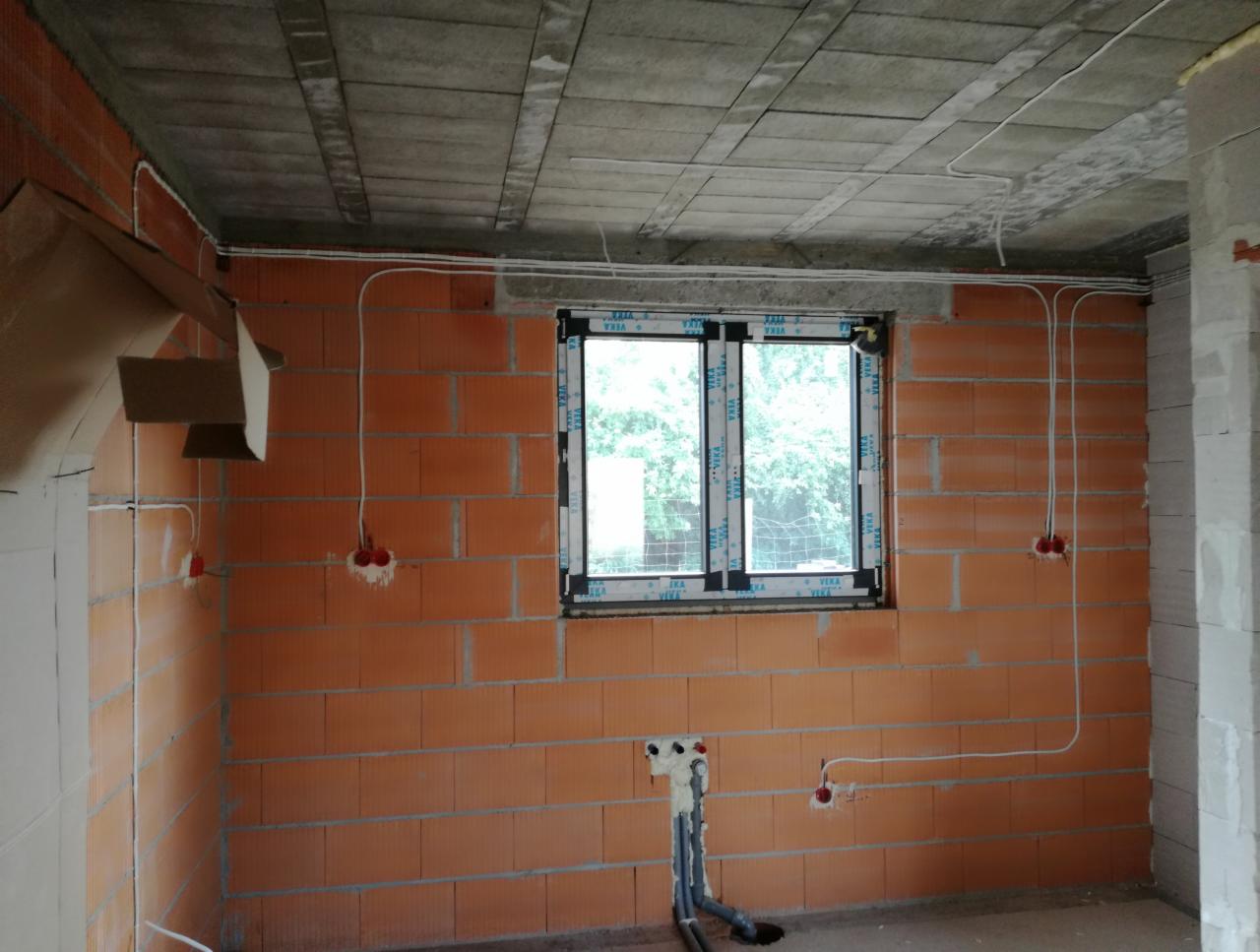 Surowy stan wnętrza z widocznymi przewodami elektrycznymi prowadzonymi po ścianach z cegły, instalacja podtynkowa w trakcie realizacji, okno z widokiem na zieleń.