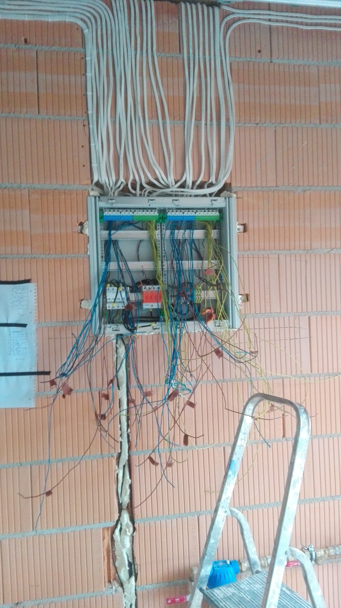 Rozdzielnica elektryczna w trakcie montażu na ceglanej ścianie, widoczne kable w peszlach oraz drabina aluminiowa z niebieską osłoną filtra wody w tle.
