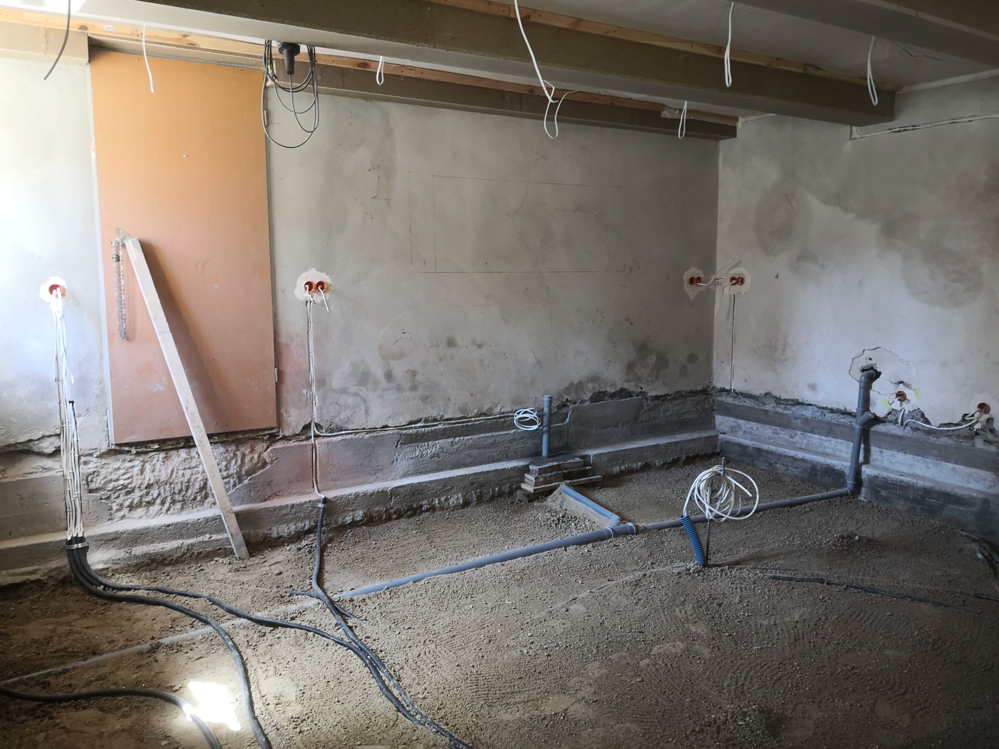 Surowy stan podłogi w pomieszczeniu z widocznymi przewodami elektrycznymi wyprowadzonymi ze ściany oraz rurami kanalizacyjnymi, przygotowanie do wylewki.
