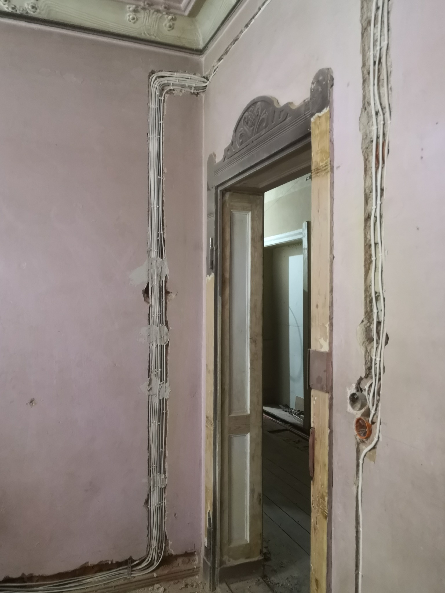 Instalacja elektryczna w starym domu: białe przewody prowadzone po powierzchni ściany, zabezpieczone gipsem, widoczne puszki instalacyjne, dekoracyjny sufit i futryna drzwi.
