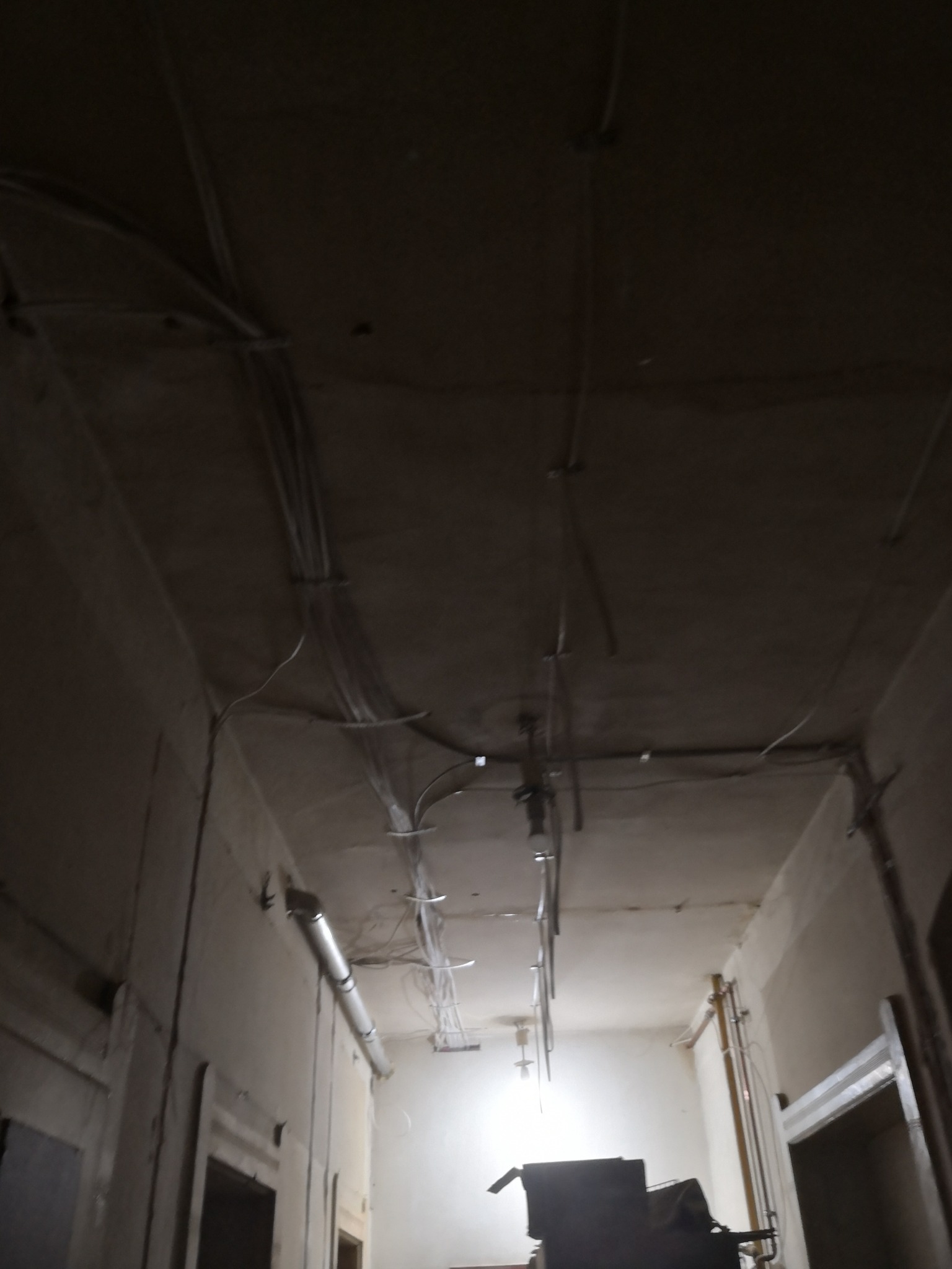 Wnętrze w trakcie remontu z widocznymi przewodami elektrycznymi prowadzonymi po suficie i ścianach, rurami wentylacyjnymi oraz prowizorycznym oświetleniem.