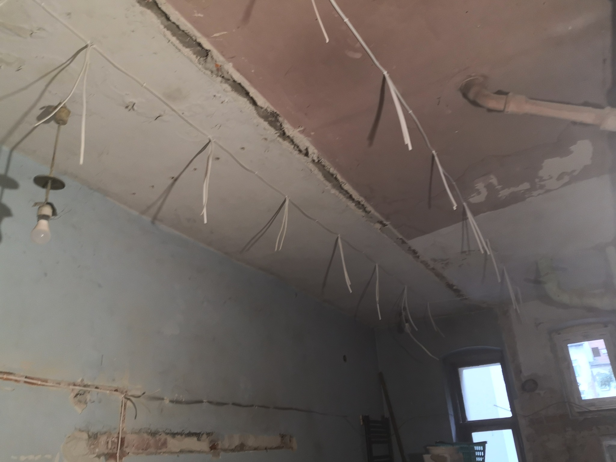 Wnętrze w trakcie remontu z widocznymi przewodami elektrycznymi zwisającymi z sufitu, stara lampa bez klosza, surowy tynk i fragment rury wentylacyjnej.