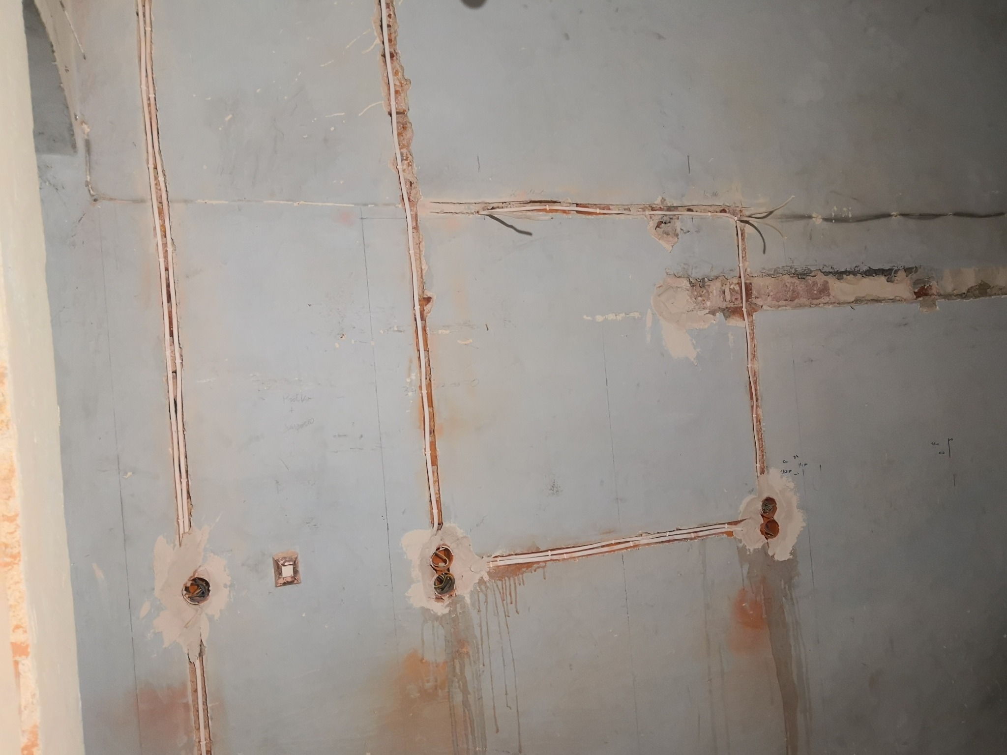 Ściana w trakcie remontu z wyżłobionymi bruzdami i przewodami elektrycznymi, widoczne puszki instalacyjne zamontowane w ścianie, szare tło ściany z widocznymi śladami po malowaniu.
