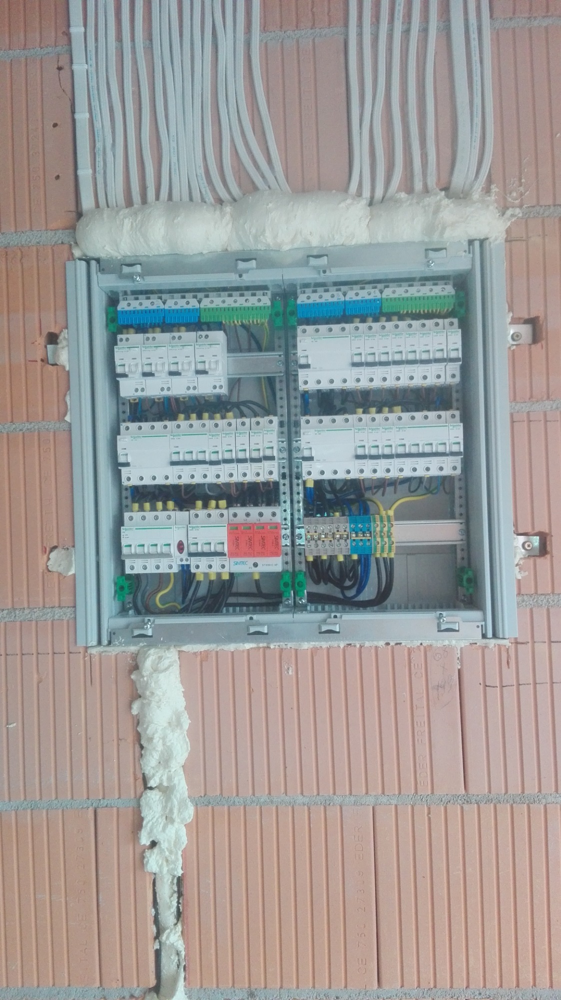 Rozdzielnica elektryczna wmurowana w ścianę z cegły, zabezpieczona pianką montażową, z widocznymi przewodami i elementami instalacji.
