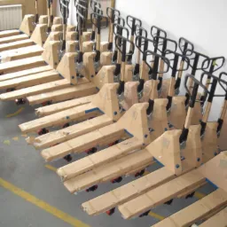 Rząd nowych, zapakowanych w karton wózków paletowych ręcznych z czarnymi uchwytami, ustawionych w magazynie na tle białej ściany i okna.