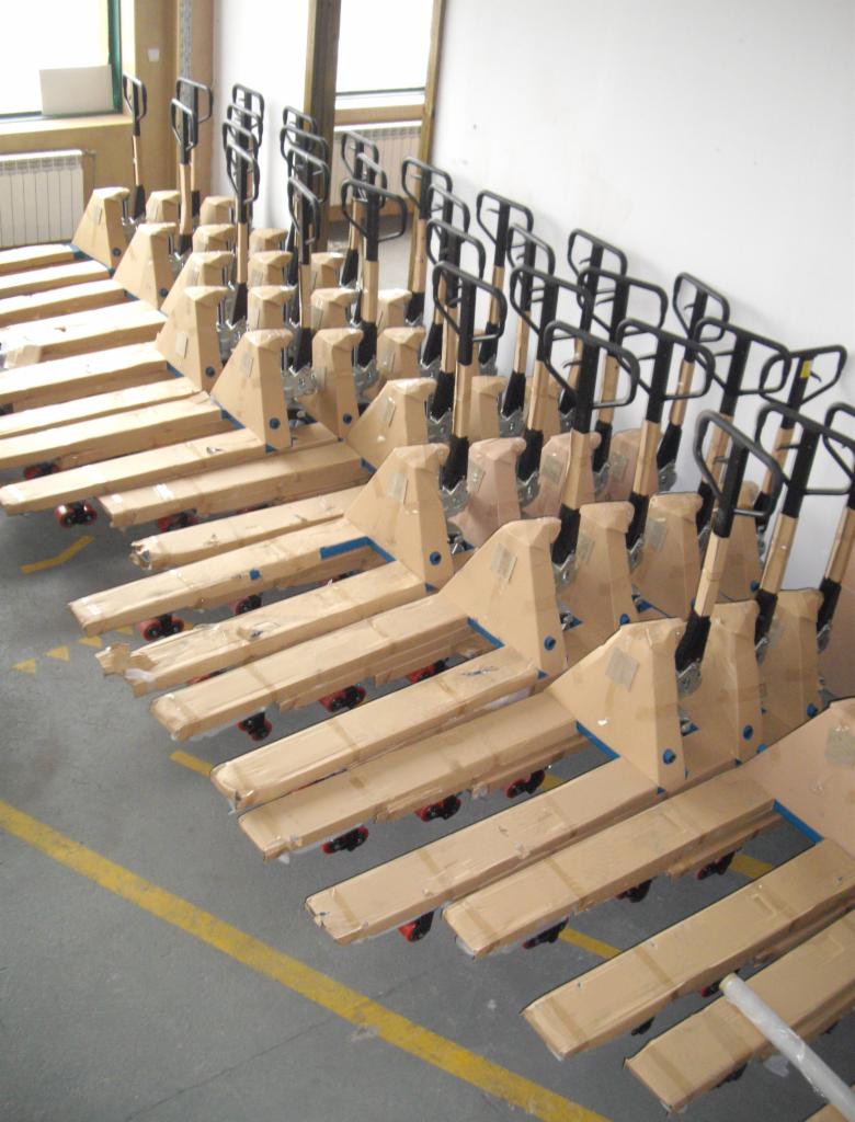 Rząd nowych, zapakowanych w karton wózków paletowych ręcznych z czarnymi uchwytami, ustawionych w magazynie na tle białej ściany i okna.