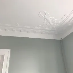 Jasnoszara ściana z białą listwą przysufitową i dekoracyjnym sztukateryjnym ornamentem na suficie, przedstawiającym postać kobiecą.