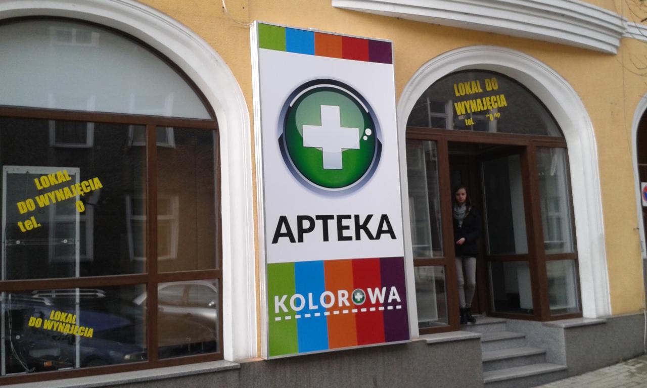 Szyld apteki Kolorowa z zielonym krzyżem w okręgu i kolorowymi paskami na górze i dole, umieszczony na fasadzie budynku z oknami z napisem 'Lokal do wynajęcia'.